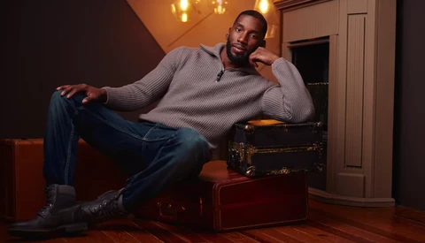 Man in gray sweater and jeans reclining on vintage luggage in warm interior lighting.