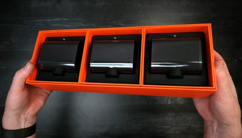 Person holding an orange display case containing three black square lens filters or optical elements.