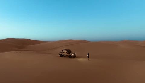 A rugged vehicle and lone figure on vast, undulating sand dunes under clear blue sky.