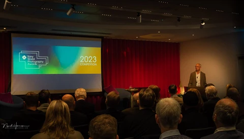 Conference speaker presenting to seated audience in auditorium with projection screen displaying 2023 event branding.