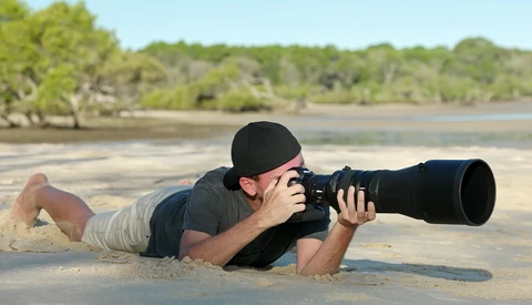 Photographer lying prone on sand while shooting with a telephoto lens toward mangrove trees.