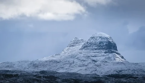 Snow-covered mountain peak with distinctive layered rock formations against a blue sky with clouds.