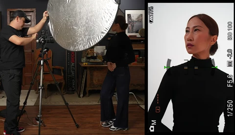 Studio portrait setup with large reflector on tripod and subject in black clothing against white background.