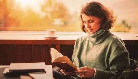 Woman in gray turtleneck reading a book by a sunlit window with coffee cup nearby.
