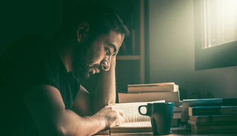 Man reading an open book at a desk with stacked books and a cup of coffee, lit by warm window light.