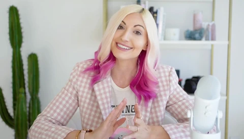 Woman with blonde and magenta hair wearing a pink gingham blazer, smiling at camera in bright interior space.
