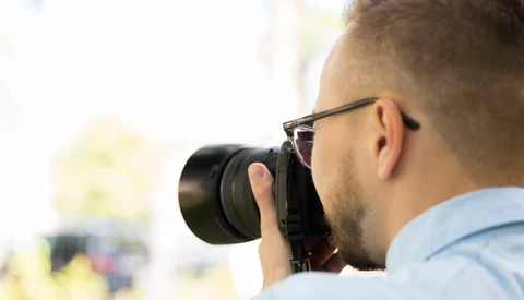 Photographer wearing glasses and light blue shirt holding camera to face while shooting outdoors.