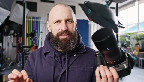 Bearded photographer holding a professional DSLR camera in a studio setting with lighting equipment visible.