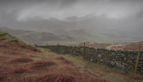 Misty hillside landscape with stone wall and moorland under overcast sky.