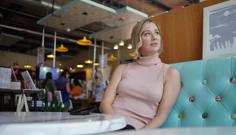 Woman in pink sleeveless turtleneck sitting on turquoise couch in modern office space.