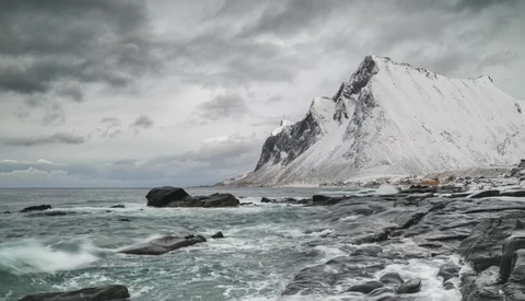 Snow-covered mountain peak rising from rocky shoreline with turbulent sea and overcast sky.