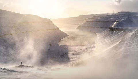 Solitary figure standing in misty coastal valley between towering cliff faces at sunrise or sunset.