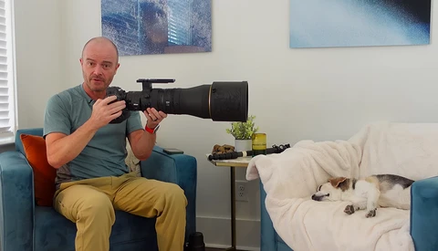 Photographer with telephoto lens posing on couch next to cat on bed.