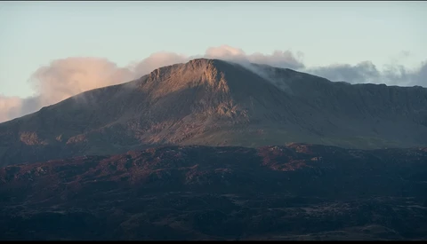Volcanic mountain peak with layered rock formations bathed in golden morning light beneath clearing clouds.