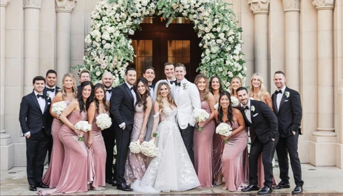 Bride and groom centered with wedding party in formal attire arranged in front of classical columns and floral archway.