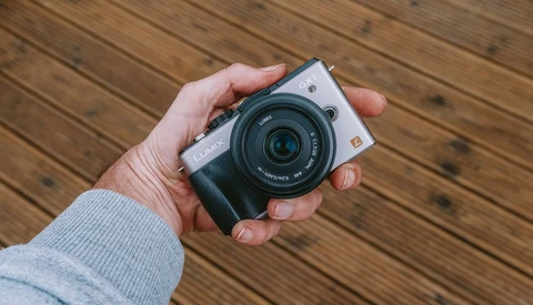 Hand holding a compact mirrorless camera with a fixed lens against a wooden surface.