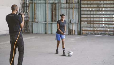 Photographer with camera positioned left, shooting a male athlete in athletic wear posing with a soccer ball in an industrial concrete space.