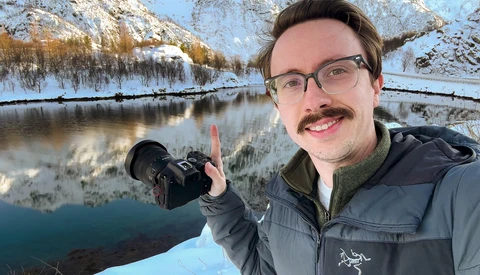 Photographer holding a camera with snow-covered mountains and a frozen river in the background.