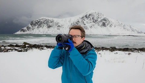 Photographer in blue jacket holding telephoto lens toward snow-covered mountain across water.