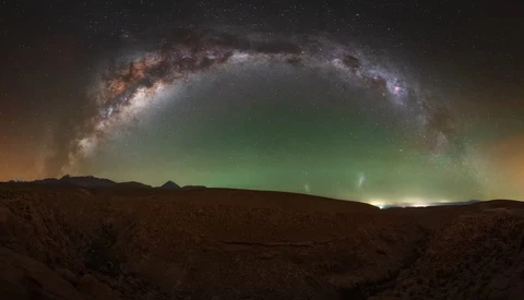 Milky Way arching over a desert landscape with green aurora lights on the horizon.