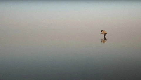 Person standing alone in shallow water with mirror-like reflections under overcast sky.