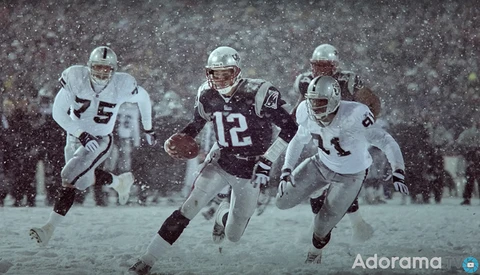 Football players in heavy snow, one in dark uniform carrying the ball while two in white uniforms defend.