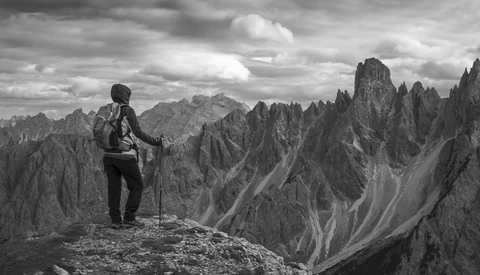 Hiker with backpack and trekking poles standing on a rocky ridge overlooking dramatic jagged peaks.