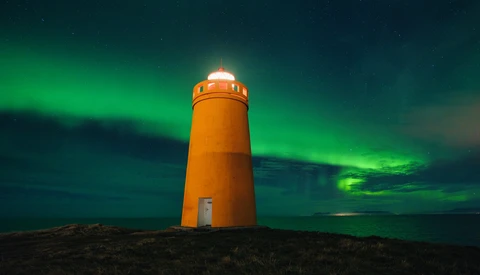 Bright orange lighthouse standing alone on dark terrain beneath vibrant green aurora borealis.