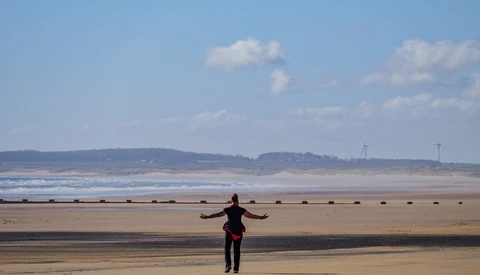 Person with arms outstretched standing alone on a wide, wet sandy beach with gentle waves and distant hills.