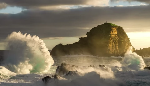 Massive waves crash against a rocky coastal outcrop during golden hour, with dramatic cloudy sky overhead.
