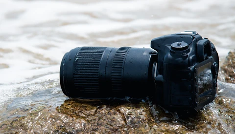 Black DSLR camera with telephoto lens partially submerged in shallow ocean water on a rocky shore.