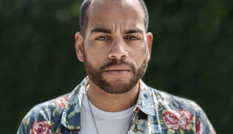 Man with beard wearing floral patterned shirt and white undershirt, photographed outdoors with blurred green background.