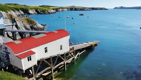 Red-roofed boathouse on wooden stilts overlooking a turquoise cove with white cliffs and moored boats.