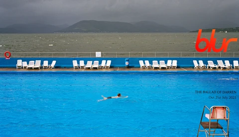 Swimmer in turquoise pool with white loungers and gray sea beyond. Mountains visible across the water.
