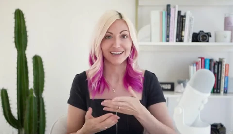 Woman with blonde and pink ombre hair wearing black shirt, gesturing while speaking to camera in bright home office.