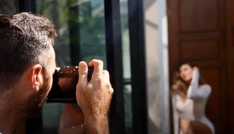 Photographer holding a camera up to his face, capturing an image of a woman visible through a doorway in the background.