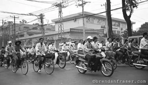 Large group of people on bicycles and motorcycles gathered on a wide street in an urban area.