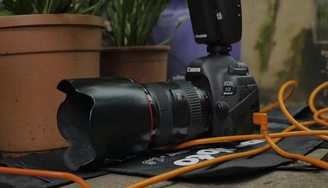 Canon EOS camera with telephoto lens and external flash on orange cables next to potted plants.