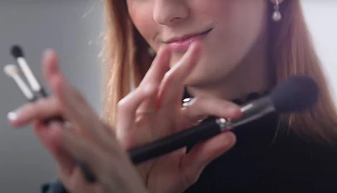 Woman with reddish-brown hair holding makeup brushes and examining cosmetic products near her face.