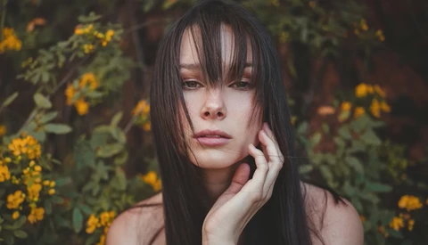 Woman with long dark hair and bangs gazing pensively beside blooming yellow flowers.