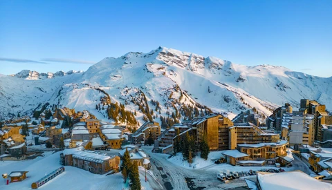 Snow-covered alpine resort nestled below a dramatic snow-capped mountain peak under clear blue sky.