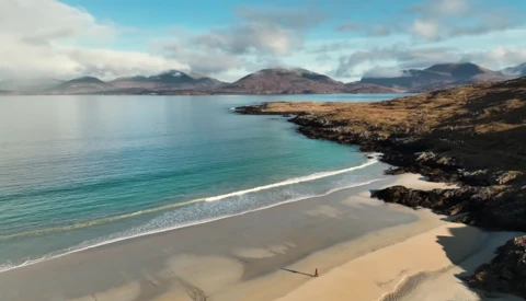 Coastal beach with turquoise water, golden sand, and dark rocky outcrops under blue sky with mountains in distance.
