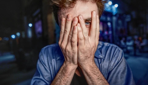 Man with reddish-brown hair covering his face with both hands, wearing a blue shirt against a blurred urban night background.