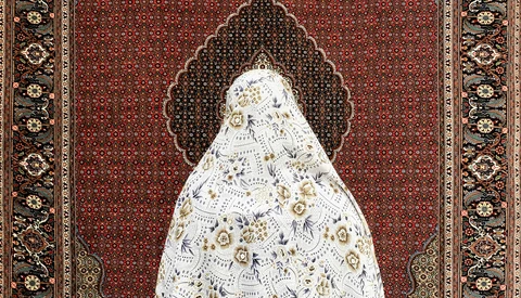 Woman in white patterned garment standing before ornate burgundy and gold traditional textile backdrop.