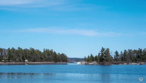 Calm blue lake with forested shoreline and boats under clear sky.