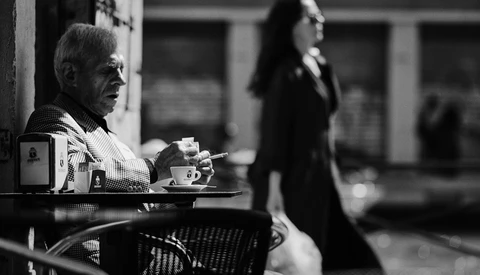 Older man in checkered shirt sitting at outdoor café table, watching passersby in black and white.