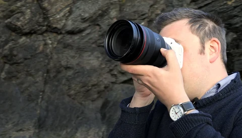 Photographer holding a telephoto lens up to his face against a rough stone wall background.