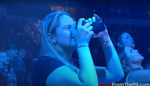 Woman singing into microphone at concert with blue stage lighting and crowd in background.