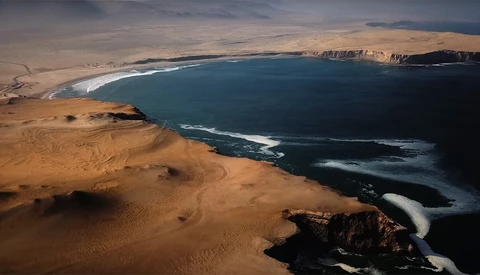 Aerial view of a vast desert coastline meeting a deep blue ocean with white surf patterns.
