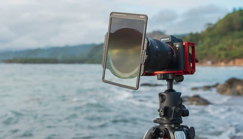 Camera mounted on tripod with smartphone holder and polarizing filter, aimed at coastal landscape with mountains and forested shoreline.